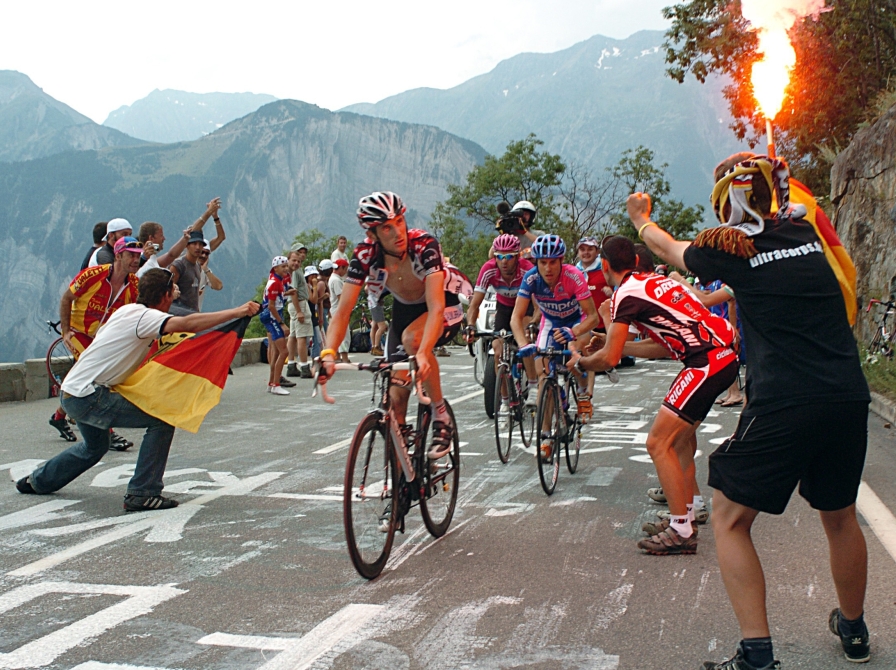 Cykelrytteren Fränk Schleck cykler på Alpe d'Huez under Tour de France 2006. Til højre ses jublende tilskuere. Én af dem med et tændt romerlys i hånden. Bag Schleck ses yderligere cykelryttere - Damiano Cunego og Eddy Mazzoleni.